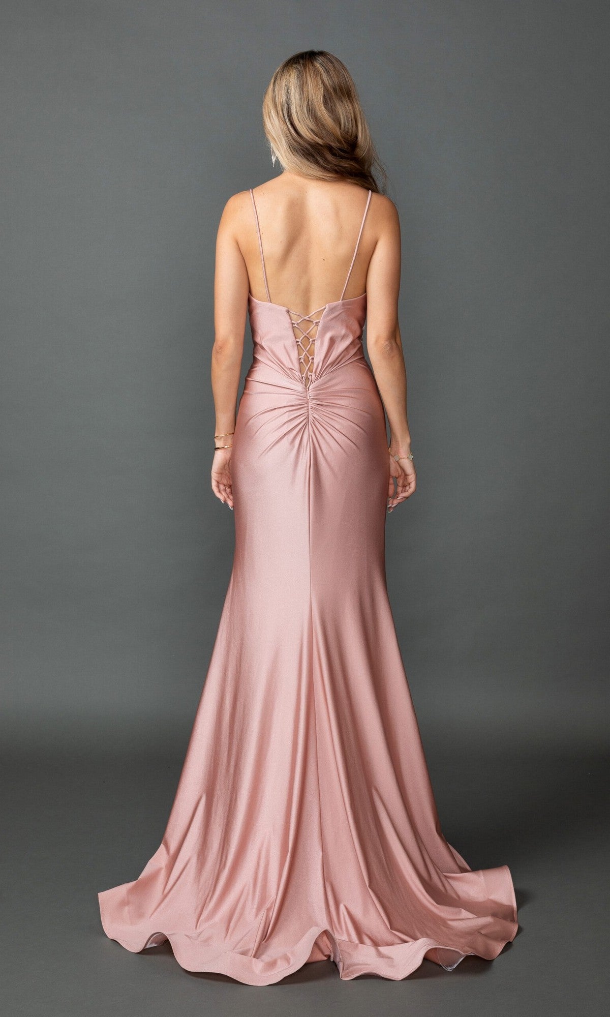A woman models the Lace-Up Long Pink Prom Dress: Dancing Queen 4553—a floor-length blush satin gown with a twist-front, thin straps, and lace-up open back—against a plain gray background.